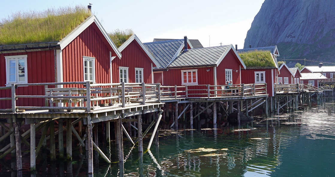 Das Bild zeigt eine Reihe von roten Holzhäusern auf den Lofoten, die auf Stelzen über dem Wasser gebaut wurden, wahrscheinlich in einem Küstengebiet. Die Häuser haben weiß gerahmte Fenster und einige haben grasbewachsene Dächer. Ein Holzsteg verbindet die Häuser, der parallel zum Wasser verläuft. Im Hintergrund ist ein großer felsiger Berg zu sehen. Die Spiegelung der Häuser und des Berges ist im Wasser darunter sichtbar. 