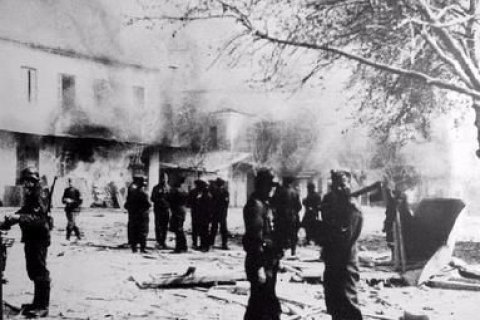 Mehrere uniformierte SS-Soldaten stehen zwischen brennenden Gebäuden und Trümmern im Dorf Distomo. Rauch steigt in den Himmel, im Vordergrund liegen zerstörte Gegenstände auf dem Boden. Kahl gewordene Bäume deuten auf Sommerhitze und Verwüstung hin. Das Foto zeigt die unmittelbaren Folgen des Massakers, das die SS am 10. Juni 1944 an der Zivilbevölkerung verübte.