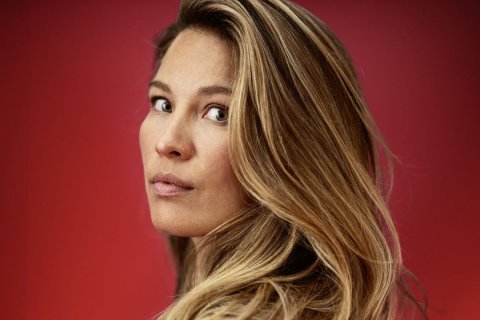 Eine Frau mit langen, leicht gewellten, blonden Haaren blickt über ihre Schulter direkt in die Kamera. Ihr Gesicht ist ernst, die Lippen geschlossen. Der Hintergrund ist einfarbig rot, was ihr Gesicht und die Haarfarbe besonders betont. Das Licht ist weich und gleichmäßig.