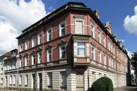 Das Bild zeigt ein großes, dreistöckiges Eckgebäude mit Mansardendach. Das Gebäude ist aus rotem Backstein gebaut und verfügt über dekorative Steinelemente an den Fenstern und entlang der Fassade. Die Fenster sind symmetrisch angeordnet, wobei jedes Stockwerk eine Reihe von rechteckigen Fenstern hat, die von Steinstürzen und Fensterbänken eingerahmt sind. Das Erdgeschoss hat eine hellere Steinfassade, die einen Kontrast zu den roten Ziegeln darüber bildet. Ein kleiner, abgerundeter Busch befindet sich an der Ecke des Gebäudes, angrenzend an den Bürgersteig. Die Architektur des Gebäudes erinnert an einen historischen Stil mit kunstvollen Details und einer prominenten Ecklage.