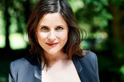 Das Bild zeigt eine Person mit schulterlangen braunen Haaren, die einen dunklen Blazer über einem hellen Oberteil trägt. Der Hintergrund besteht aus einer natürlichen Umgebung mit verschwommenem Grün, das auf das Vorhandensein von Bäumen oder Laub hinweist.