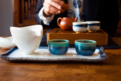 Bild eines chinesischen Teegedecks - bestehend aus einer weißen Kanne und zwei kleinen Tassen auf einem Keramiktablett - auf einem Holztisch; im Hintergrund sieht man die Hand einer Person, die den Deckel einer weiteren Keramik-Teekanne öffnet, neben der zwei weitere Teetassen stehen.