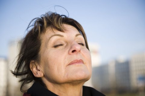 Im Vordergrund ist der Kopf einer Frau vor blauem Himmel und unscharf gehaltenen Gebäuden im Hintergrund zu sehen. Die Frau scheint den Kopf in den Wind zu halten. Die Haare sind ein wenig zerzaust, sie hat Augen und Mund geschlossen und scheint durch die Nase tief durchzuatmen. 