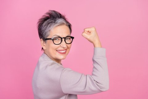 Vor einem pinken Hintergrund ist eine Frau mittleren Alters mit Kurzhaarfrisur, Brille und im Blazer zu sehen, die ihren rechten Arm angewinkelt hält und eine Faust macht, als würde sie ihren Bizeps zeigen. Sie lächelt energiegeladen und zuversichtlich in die Kamera. 