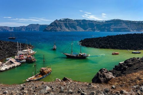 Das Bild zeigt eine K&uuml;stenszene mit mehreren Booten, die in einer Bucht positioniert sind, die von felsigem Gel&auml;nde umgeben ist.

1. **Vordergrund**: Der Vordergrund besteht aus felsigem Untergrund mit sichtbaren Steinen und sp&auml;rlicher Vegetation. Die Steine sind ungleichm&auml;&szlig;ig verteilt und f&uuml;hren nach unten in Richtung Wasser.

2. **Boote**:
- Auf der linken Seite des Bildes sind mehrere Boote zu sehen, die nahe am Ufer angedockt sind. Diese Boote sind gr&ouml;&szlig;er, mit sichtbaren Masten und Strukturen, die an traditionelle h&ouml;lzerne Segelschiffe erinnern.
- In der Mitte der Bucht befindet sich ein einzelnes Boot, das weiter vom Ufer entfernt positioniert ist und mit einem Mast und Segeln mittelgro&szlig; aussieht.
- Auf der rechten Seite der Bucht gibt es kleinere Boote, darunter eines, das wie ein Motorboot aussieht. Diese Boote befinden sich n&auml;her am Felsvorsprung auf der rechten Seite.

3. **Wasser**: Das Wasser in der Bucht wechselt von einem gr&uuml;nlichen Farbton in der N&auml;he des Ufers zu einem tieferen Blau weiter drau&szlig;en. Die gr&uuml;ne F&auml;rbung konzentriert sich auf die Boote und die felsige K&uuml;ste.

4. **Felsvorsprung**: Auf der rechten Seite des Bildes befindet sich ein gro&szlig;er, dunkler Felsvorsprung, der ins Wasser ragt. Dieser Aufschluss trennt das gr&uuml;nliche Wasser vom tieferen blauen Wasser dahinter.

5. **Hintergrund**: Der Hintergrund zeigt eine gro&szlig;e Klippe oder eine bergige Formation, die sich &uuml;ber die gesamte Breite des Bildes erstreckt. Die Klippen sind steil und zerkl&uuml;ftet, wobei einige Bereiche oben Schnee oder hellere Flecken zu haben scheinen. Die Klippen sind quer &uuml;ber die Bucht positioniert und bilden eine nat&uuml;rliche Grenze.

6. **Himmel**: &Uuml;ber den Klippen ist der Himmel mit vereinzelten Wolken zu sehen. Der Himmel nimmt den oberen Teil des Bildes ein.