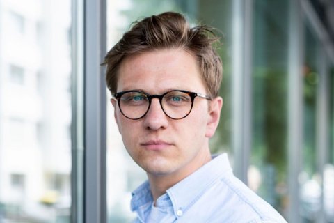 Portr&auml;taufnahme eines jungen Mannes mit hellbraunem, nach oben gestyltem Haar und runder Schildpatt-Brille mit blauen Gl&auml;sern. Er blickt direkt in die Kamera, der Gesichtsausdruck ist neutral. Er tr&auml;gt ein hellblaues Hemd. Der Hintergrund zeigt eine Glasfassade, dahinter Geb&auml;ude und Gr&uuml;n, unscharf. Foto: Elisa Prod&ouml;hl