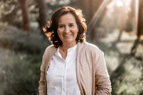 Frau mit braunen Haaren, wei&szlig;er Bluse und Strickjacke, die gl&uuml;cklich zu sein scheint, auf Waldlichtung; Sonnenlicht f&auml;llt seitlich von hinten ein und scheint durch die Haare