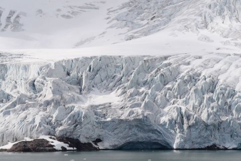 Ein m&auml;chtiger Gletscher mit zackigen, blau-wei&szlig;en Eismassen schiebt sich ins Meer. Die Oberfl&auml;che zeigt Risse, Spalten und gefaltete Strukturen. Unten erkennt man eine dunkle Felsbasis, an der das Eis direkt ins Wasser m&uuml;ndet. Das Meer im Vordergrund ist ruhig, mit wenigen kleinen Eisschollen.