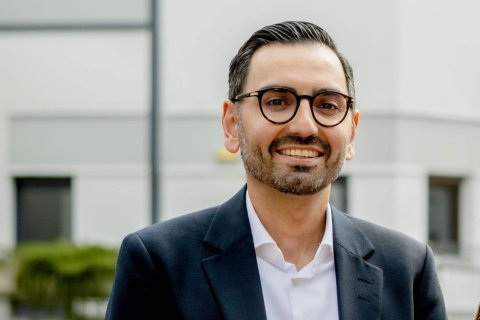 Portr&auml;taufnahme eines etwa 40-j&auml;hrigen Mannes mit kurzen, nach hinten gek&auml;mmten dunklen Haaren und gepflegtem Vollbart. Er tr&auml;gt eine runde Brille mit dunklem Horngestell, ein dunkelblaues Sakko und ein wei&szlig;es, am Kragen offenes Hemd. Er blickt l&auml;chelnd in die Kamera, die Z&auml;hne sind sichtbar. Der Bildausschnitt reicht von der Brust aufw&auml;rts. Im unscharfen Hintergrund eine helle, moderne Geb&auml;udefassade mit grauen Fensterb&auml;ndern, links unten ein gr&uuml;ner Strauch.