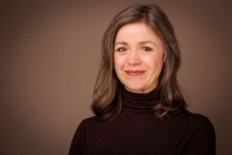 Portr&auml;t einer Frau mittleren Alters vor braunem Studiohintergrund. Sie blickt mit geschlossenem L&auml;cheln in die Kamera. Ihr schulterlanges braunes Haar mit grauen Str&auml;hnen f&auml;llt locker &uuml;ber die Schultern. Sie hat braune Augen und tr&auml;gt dezenten dunkelroten Lippenstift sowie einen dunkelbraunen Rollkragenpullover. Weiches Licht f&auml;llt von vorn auf das Gesicht, der Hintergrund verl&auml;uft von hellem zu dunklem Braun.