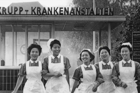 Schwarz-Wei&szlig;-Aufnahme, vermutlich 1960er Jahre. F&uuml;nf junge Frauen in Krankenschwesternuniformen &ndash; helles Oberteil, wei&szlig;es Latzsch&uuml;rze, wei&szlig;e Haube &ndash; stehen nebeneinander vor einem Flachdachgeb&auml;ude. Alle l&auml;cheln. Im Hintergrund gro&szlig;er Schriftzug: &bdquo;KRUPP-KRANKENANSTALTEN". Rechts ein Hinweisschild mit Abteilungsnamen, teils lesbar.