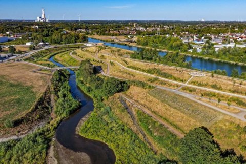 Luftaufnahme einer Flusslandschaft im Sommerlicht. Im Vordergrund windet sich ein schmaler, dunkler Fluss mit Kiesufern durch gr&uuml;ne B&ouml;schungen. Rechts daneben steigen Wiesenterrassen mit hellen Wegen an. In das trockene Gras ist in gro&szlig;en Buchstaben &bdquo;EmscherLand&ldquo; geschrieben. Dahinter verl&auml;uft ein breiter, blauer Kanal, den eine geschwungene Br&uuml;cke &uuml;berspannt. Am Horizont liegen Wohnsiedlungen, W&auml;lder, Windr&auml;der und links ein Kraftwerk mit Dampfwolke unter klarem Himmel.