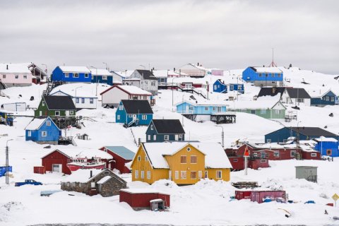 Mehrere bunte Holzhäuser in kräftigen Farben wie Gelb, Blau, Grün und Rot stehen auf verschneiten Hängen. Schneebedeckte Dächer, Strommasten und ein Boot im Vordergrund prägen die winterliche Szenerie unter grauem Himmel.