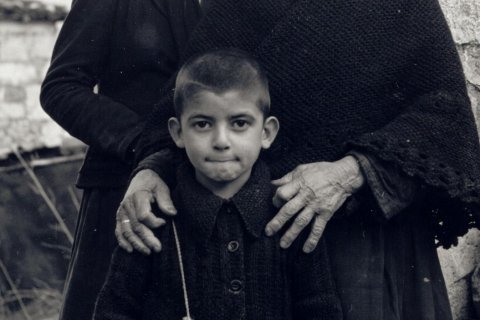 Das Bild zeigt drei Individuen. Im Vordergrund ist ein Kind zu sehen. Das Kind trägt ein zugeknöpftes Kleidungsstück mit sichtbarem Kragen. Hinter dem Kind sind zwei Erwachsene anwesend. Der Erwachsene auf der linken Seite trägt ein dunkles, langärmeliges Kleidungsstück, der linke Arm ruht auf der Schulter des Kindes. Der Erwachsene auf der rechten Seite trägt einen strukturierten Schal oder ein Tuch, die Hände ruhen auf den Schultern des Kindes. Die Hände des Erwachsenen auf der rechten Seite sind deutlich zu sehen und zeigen Altersspuren. Im Hintergrund gibt es eine Steinmauer und einige undeutliche Elemente.