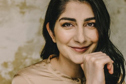 Die Journalistin Waslat Hasrat-Nazimi, Foto: Julia Sellmann Photography Das Bild zeigt eine Person mit langen, dunklen Haaren, die über die rechte Schulter fallen. Sie trägt eine glänzende, beige Bluse mit kleinem Stehkragen und Knopf am Ausschnitt. Eine Hand ist angewinkelt; die Finger ruhen locker unter dem Kinn. Die Person blickt direkt in die Kamera, die Lippen formen ein leises Lächeln. Der Hintergrund wirkt texturiert in creme- und ockerfarbenen Tönen.