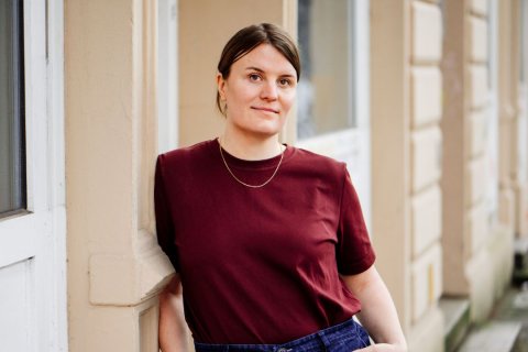 Das Bild zeigt eine Person, die ein kastanienbraunes kurzärmeliges Hemd und eine dünne Goldkette trägt. Das Shirt hat ein schlichtes Design ohne sichtbare Muster oder Logos. Die Person trägt auch eine blaue Jeanshose. Ihr linker Arm ruht auf einer hellen vertikalen Fläche, möglicherweise einer Wand oder Säule. Der Hintergrund enthält eine verschwommene Ansicht von etwas, das wie ein Fenster oder eine Glasscheibe mit vertikalem Rahmen aussieht. Das Haar der Person ist dunkel und zurückgebunden.