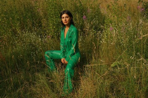 Eine Frau mit dunklem, kinnlangem Haar sitzt auf einer sommerlichen Wiese. Sie tr&auml;gt einen gl&auml;nzenden, smaragdgr&uuml;nen Hosenanzug mit tiefem V-Ausschnitt. Ihre H&auml;nde ruhen locker auf den Oberschenkeln, der Blick geht direkt in die Kamera. Das Gras reicht ihr bis zur H&uuml;fte, dazwischen wachsen wei&szlig;e und violette Wildblumen. Warmes, goldenes Licht f&auml;llt von links auf die Szene.