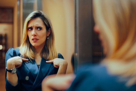 Eine Frau mit langen Haaren, Ohrring und einem Blazer betrachtet sich selbst zweifelnd im Spiegel. Wir sehen ihr Spiegelbild scharf und frontal, sie selbst nur unscharf von hinten. Ihre Stirn ist gekräuselt, der Blick unsicher und sie zeigt fragend mit beiden Zeigefingern auf sich selbst, als wollte sie sagen: "Kann ich das? Darf ich das? Bin ich das?"