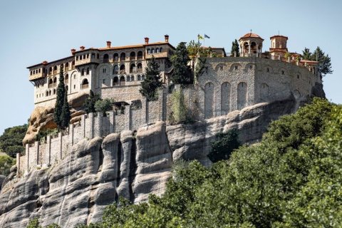 Auf einem hohen, steil aufragenden Felsen steht eine gro&szlig;e orthodoxe Klosteranlage aus hellem Stein. Mehrere Geb&auml;ude mit roten Ziegeld&auml;chern schmiegen sich an den Felsrand. Eine massive St&uuml;tzmauer folgt der nat&uuml;rlichen Form des Gesteins. Im Vordergrund wachsen gr&uuml;ne B&auml;ume, dar&uuml;ber spannt sich ein klarer, hellblauer Himmel.
