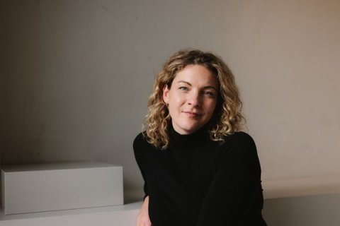 Eine Frau mit hellen, lockigen Haaren sitzt leicht seitlich vor einer hellen Wand. Sie tr&auml;gt einen schwarzen Rollkragenpullover und st&uuml;tzt den rechten Arm auf einen wei&szlig;en Quader. Weiches Licht f&auml;llt von links auf ihr Gesicht, ihre Miene ist offen und ruhig, der Hintergrund schlicht und leer.