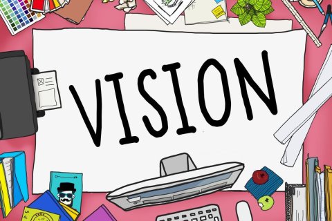 Cartoon-Darstellung einer Schreibtischoberfläche, auf der viele Dinge ausgebreitet liegen - u.a. Briefumschläge, Stifte, ein Lineal, ein Farbfächer, Bücher, ein Monitor, eine Tastatur und ein Drucker. In der Mitte liegt jedoch ein großes weißes Blatt, auf dem in schwarzen Großbuchstaben das Wort "VISION" steht. 