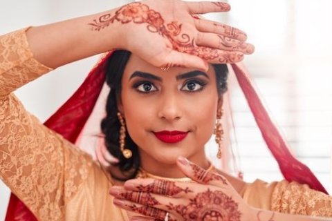 Das Bild zeigt eine Person, die ein traditionelles Outfit tr&auml;gt. Die Person tr&auml;gt ein lang&auml;rmeliges Kleidungsst&uuml;ck mit aufwendigen Spitzendetails. &Uuml;ber den Kopf ist ein roter Schleier drapiert, der sich &uuml;ber den R&uuml;cken erstreckt. Die H&auml;nde der Person sind mit aufwendigen Henna-Designs mit floralen und geometrischen Mustern geschm&uuml;ckt. Die rechte Hand wird &uuml;ber den Kopf gehoben, w&auml;hrend die linke Hand vor dem Gesicht positioniert ist. Das Henna an den H&auml;nden ist detailliert, wobei sich die Designs von den Fingern bis zu den Handgelenken erstrecken. Die Person tr&auml;gt auch goldene Ohrringe, die durch den Schleier sichtbar sind.