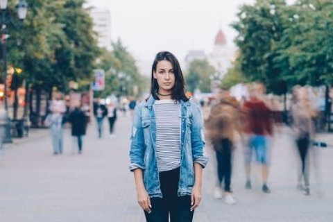 Eine Frau verweilt still inmitten vorbeieilender Passanten – Sinnbild für Innehalten im urbanen Strom. Foto: silverkblackstock/shutterstock Eine junge Frau mit langen dunklen Haaren steht auf einer belebten Straße. Sie trägt eine gestreifte Bluse, eine offene Jeansjacke und schwarze Hosen. Um sie herum gehen viele Menschen vorbei, deren Bewegungen verschwommen wirken, während sie selbst scharf und ruhig im Zentrum des Bildes bleibt. Im Hintergrund sind Bäume, Häuser und Lichter der Stadt zu erkennen.