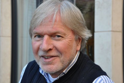 Ein &auml;lterer Mann mit grauen Haaren und kurzem Bart blickt l&auml;chelnd in die Kamera. Er tr&auml;gt ein dunkelblaues Oberteil &uuml;ber einem hellblauen Hemd mit Streifen. Im Hintergrund sind unscharf helle Steinw&auml;nde und ein Fenster zu erkennen. Das Bild ist eine Nahaufnahme, der Fokus liegt auf dem Gesicht des Mannes, das freundlich und offen wirkt.