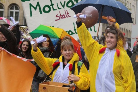 Zwei junge Frauen in gelb-wei&szlig;en H&uuml;hnerkost&uuml;men stehen im Vordergrund einer Stra&szlig;endemonstration. Beide l&auml;cheln und halten gerollte Papiere hoch. Hinter ihnen ein wei&szlig;es Transparent mit der teilweise lesbaren Aufschrift &bdquo;Motivation statt" in gr&uuml;ner und roter Schrift. Im Hintergrund weitere junge Menschen, ein regenbogenfarbener Schirm, ein schwarzer Schirm und ein brauner Luftballon. Bedeckter Himmel, urbane Geb&auml;udefassade. Orangefarbenes Banner am linken Bildrand.