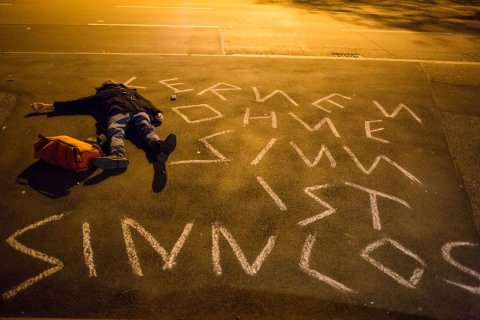 Eine Person in dunkler Jacke und Jeans liegt b&auml;uchlings auf nassem Asphalt, Arme seitlich ausgestreckt. Neben ihr ein orangefarbener Rucksack. In wei&szlig;er Kreide steht in Gro&szlig;buchstaben auf der Stra&szlig;e: &bdquo;LERNEN OHNE SINN IST SINNLOS". Die Aufnahme erfolgt aus erh&ouml;hter Perspektive. Warmes, gelblich-oranges Kunstlicht beleuchtet die Szene, der Hintergrund verschwindet in Dunkelheit. Ein St&uuml;ck Kreide liegt neben der Schrift.