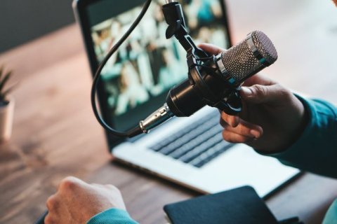 Eine Person spricht in ein Mikrofon, das an einem Schwenkarm befestigt ist. Im Hintergrund steht ein aufgeklappter Laptop mit einem Videokonferenz- oder Aufnahmebildschirm. Auf dem Tisch liegen ein Notizbuch und eine kleine Pflanze.