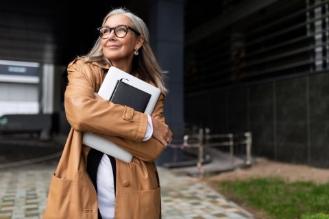 Eine Frau mit langen grauen Haaren, dunkler Brille, Lederjacke und Ohrringen steht drau&szlig;en vor einem B&uuml;rogeb&auml;ude. Sie h&auml;lt ein Laptop und ein Notizbuch in ihren Armen vor der Brust und schaut optimistisch in die Ferne - weg vom B&uuml;rogeb&auml;ude. 