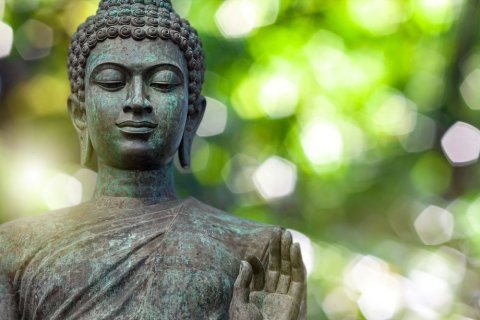 Eine bronzene Buddha-Statue mit geschlossenen Augen und ruhigem Gesichtsausdruck steht leicht seitlich vor einem unscharfen, gr&uuml;n leuchtenden Hintergrund. Die rechte Hand ist erhoben, die Finger leicht ge&ouml;ffnet &ndash; eine klassische Geste des Segens oder Schutzes. Das Lichtspiel im Hintergrund erzeugt eine friedliche, fast schwebende Atmosph&auml;re.