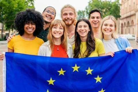 Sieben junge Erwachsene stehen dicht nebeneinander im Freien und l&auml;cheln in die Kamera. Sie halten gemeinsam eine blaue Flagge mit gelben Sternen &ndash; die Flagge der Europ&auml;ischen Union. Die Gruppe wirkt vielf&auml;ltig, ihre Kleidung ist sommerlich und farbenfroh. Im Hintergrund sind B&auml;ume und Geb&auml;ude zu sehen, die auf eine st&auml;dtische Umgebung hinweisen.