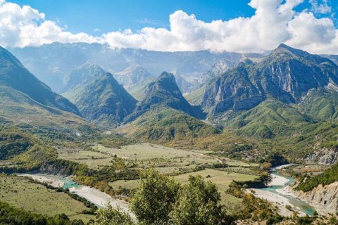 Weiter Blick &uuml;ber ein gr&uuml;nes Flusstal in Albanien. Ein t&uuml;rkisfarbener Fluss m&auml;andert in mehreren Armen durch ein breites Kiesbett, umgeben von Feldern, Baumgruppen und vereinzelten H&auml;usern. Dahinter staffeln sich bewaldete Bergketten bis zu schroffen, kahlen Gipfeln, &uuml;ber denen wei&szlig;e Wolken h&auml;ngen. Im Vordergrund die Krone eines Baumes.