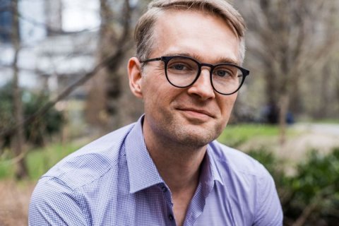Portr&auml;t von Julian Hei&szlig;ler im Freien. Der Mann mit blondem, seitlich gescheiteltem Haar blickt direkt in die Kamera und l&auml;chelt leicht. Er tr&auml;gt eine runde, dunkle Brille und ein blau-wei&szlig; kariertes Hemd mit offenem Kragen. Im unscharfen Hintergrund kahle B&auml;ume, links die Fassade eines Geb&auml;udes, rechts gr&uuml;ne Str&auml;ucher. Ged&auml;mpftes Tageslicht. Foto: Chris Sorensen